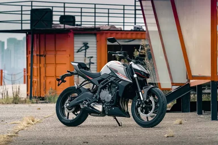 White and black KOVE naked motorcycle with red accents, parked on gravel in front of orange industrial shipping containers