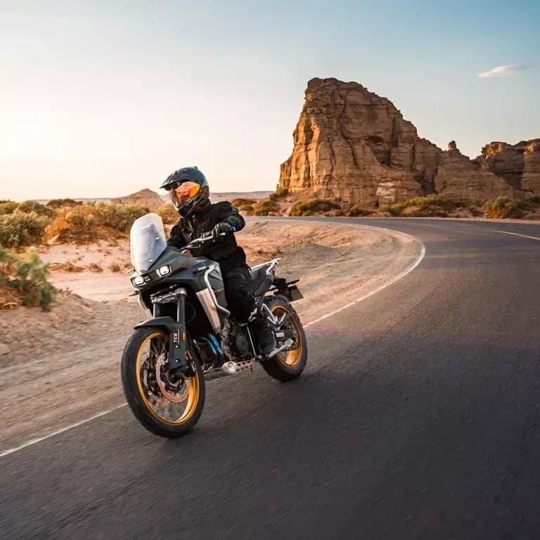 Rider in black helmet and gear riding a grey KOVE adventure motorcycle along a curving road through a rocky desert landscape at sunset
