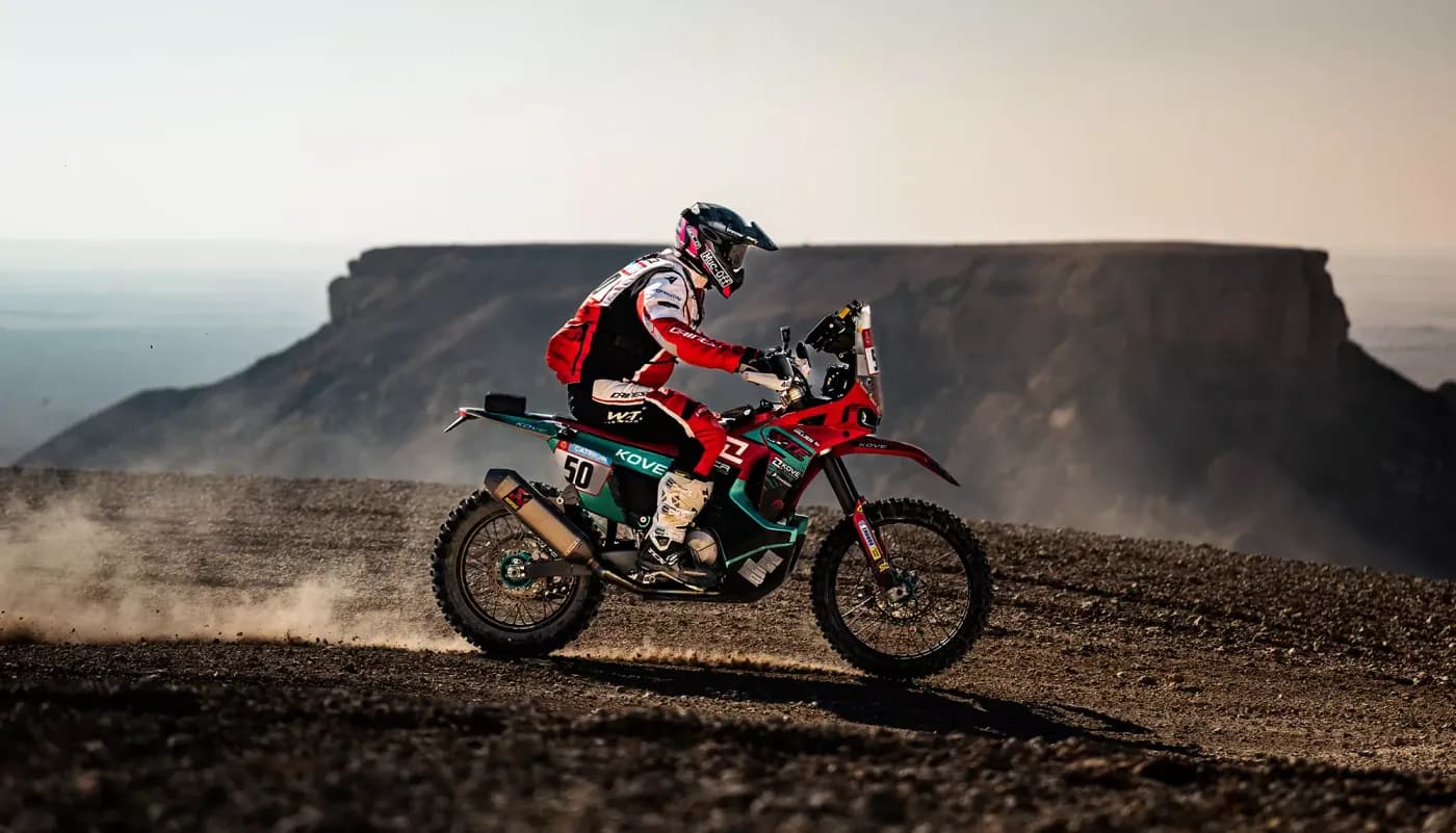 Rally rider on a red, white and teal KOVE motorcycle kicking up dust on stony terrain with a rocky plateau behind; race number 50 visible