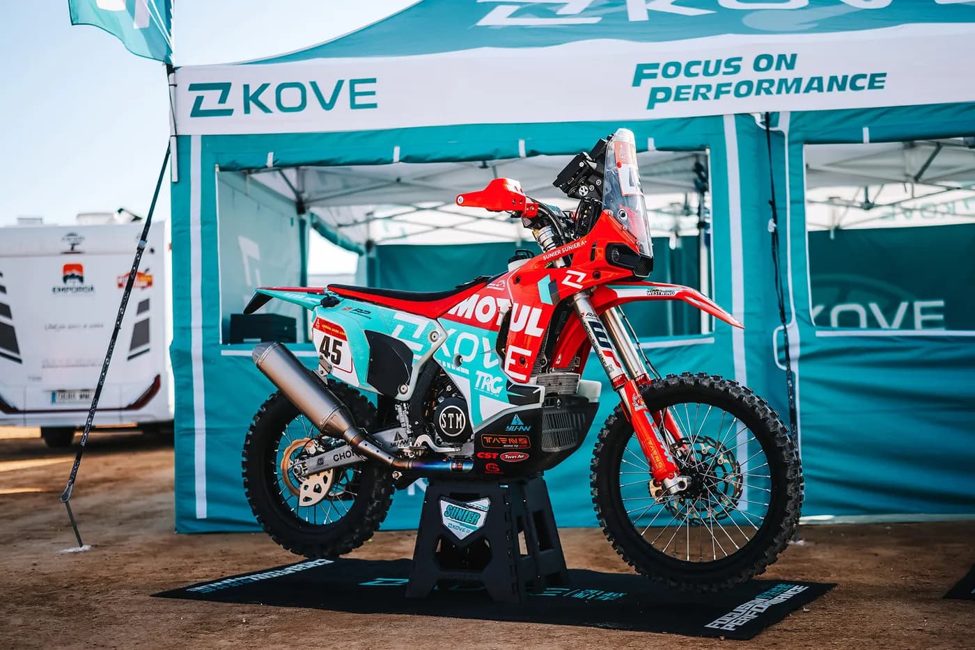 KOVE rally motorcycle on a paddock stand under a KOVE team awning reading “Focus on Performance”; race number 45 and the name “SUNIER” are legible