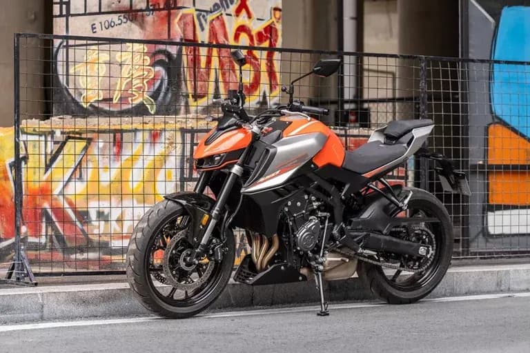 Orange and silver KOVE naked motorcycle parked at the kerb in front of a metal fence and a graffiti-covered wall