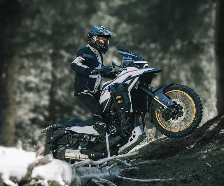 Rider in helmet and enduro jacket riding a white and blue KOVE adventure motorcycle along a dirt trail in a forest