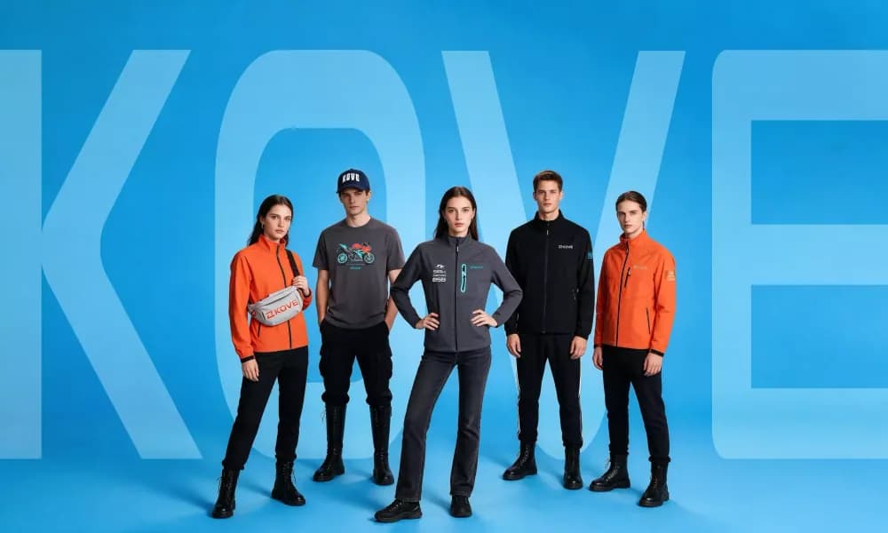 Five models against a blue backdrop with a large KOVE wordmark behind them, wearing orange and grey softshell jackets, a grey t-shirt, a black jacket, a navy cap and a grey KOVE waist bag