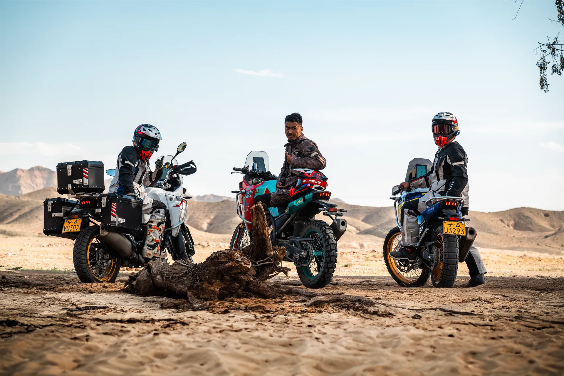 Three riders in adventure gear pause on arid ground, astride KOVE motorcycles, with mountains in the background.