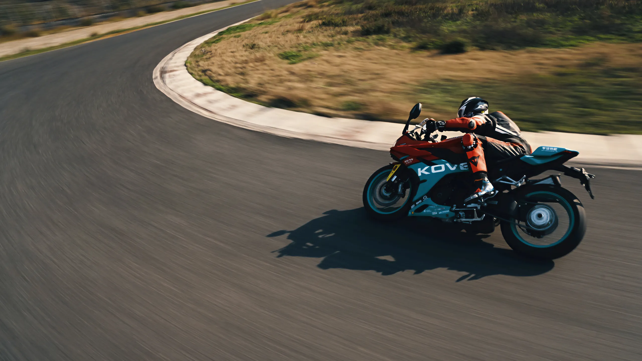 A teal-and-black KOVE sportbike with the KOVE name on its fairing, leaning into a racetrack corner; the rider wears orange leathers and the background is blurred by speed