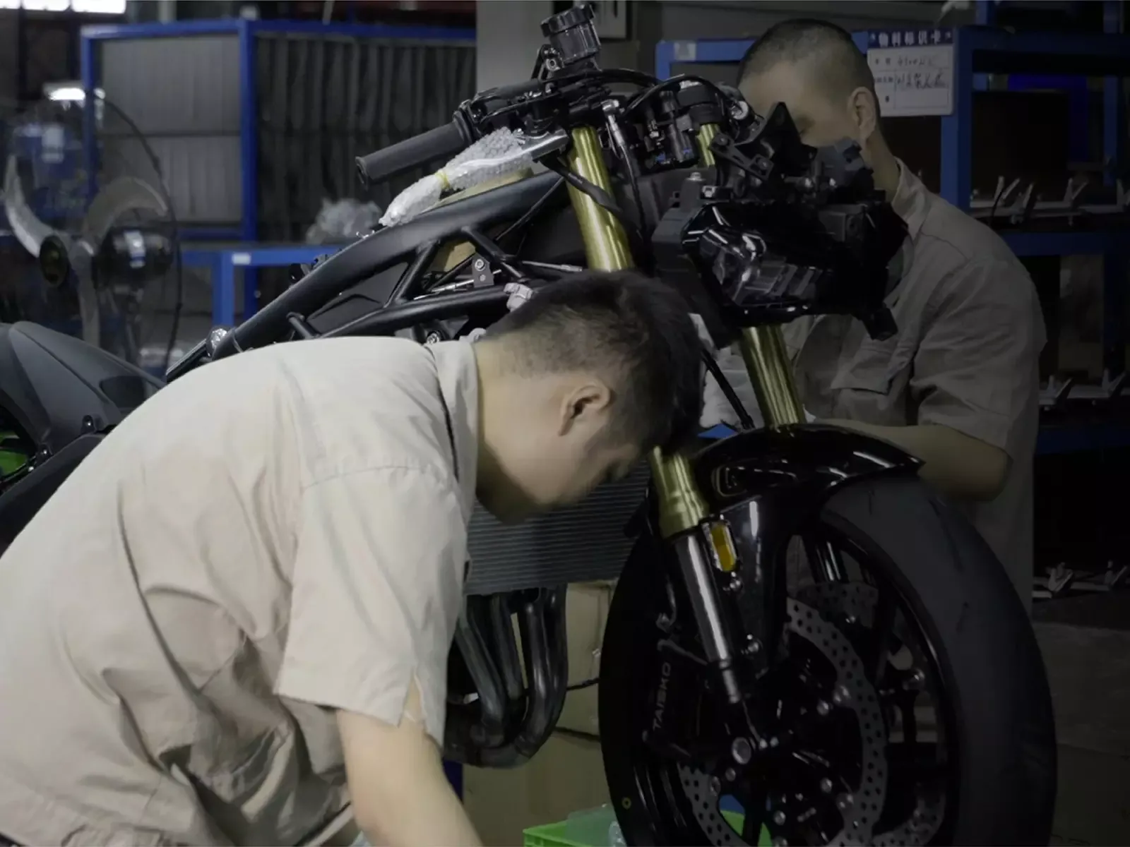 Two workers assemble a KOVE motorcycle, still unfaired, on the assembly line at the KOVE factory: the golden fork, the front brake disc and the bare frame are visible.
